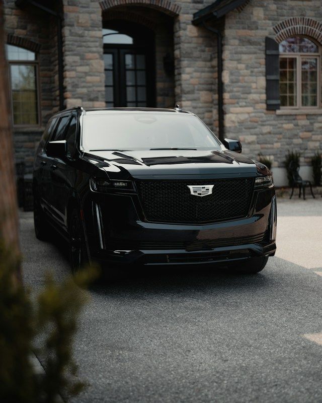 Black Cadilac Escalade in front of a large stone house.